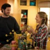 "A man and woman smile and toast with mugs in a cozy, sunlit kitchen, surrounded by yellow cabinets, kitchen appliances, and decorative items, creating a warm, homey atmosphere."