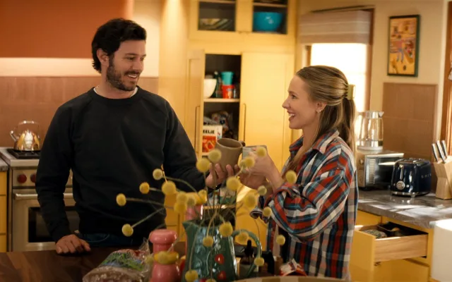 Η τρίτη σεζόν του Κανείς Δεν το Θέλει στο Netflix φέρνει τη Joanne και τον Noah κάτω από την ίδια στέγη 2 "A man and woman smile and toast with mugs in a cozy, sunlit kitchen, surrounded by yellow cabinets, kitchen appliances, and decorative items, creating a warm, homey atmosphere."