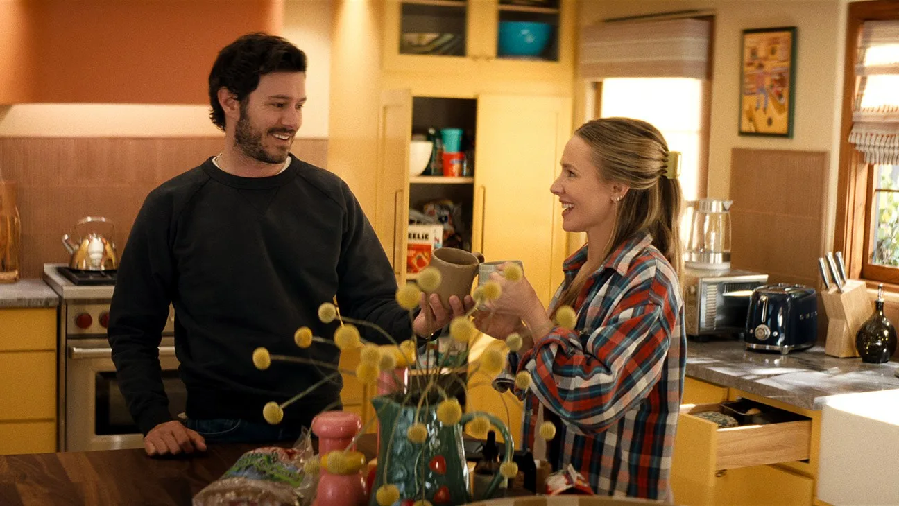 Η τρίτη σεζόν του Κανείς Δεν το Θέλει στο Netflix φέρνει τη Joanne και τον Noah κάτω από την ίδια στέγη 1 "A man and woman smile and toast with mugs in a cozy, sunlit kitchen, surrounded by yellow cabinets, kitchen appliances, and decorative items, creating a warm, homey atmosphere."